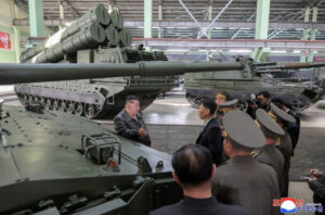 officials and soldiers listen to a briefing beside a massive battle tank with a long cannon inside a warehouse